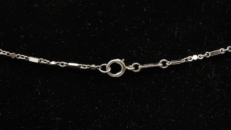 May include: Close-up of a silver necklace clasp and chain against a black background. The chain features a delicate design with small, rectangular links and circular connectors. The clasp is a simple, secure design.