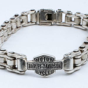 May include: A silver Harley-Davidson motorcycle chain bracelet with a silver logo charm that reads "MOTOR HARLEY-DAVIDSON CYCLES".