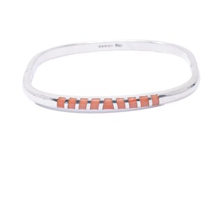 Peut inclure: Un bracelet jonc articulé de couleur argentée orné d'une rangée de petites incrustations rectangulaires de couleur corail sur la partie avant. Le bracelet présente une finition métallique lisse et polie ainsi qu'une forme ovale légèrement carrée.