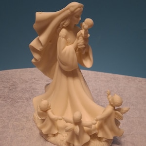 Op de afbeelding: Ivoor kleurige beeldje dat een vrouw met een baby afbeeldt, omringd door cherubijnen. De vrouw draagt een lange jurk en sluier, met cherubijnen die elkaars handen vasthouden in een cirkel. Het beeldje is gedetailleerd en staat op een voet.