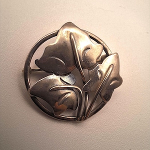 May include: A silver brooch with a circular design featuring three stylized leaves. The leaves are detailed with veins and are arranged in a cluster.