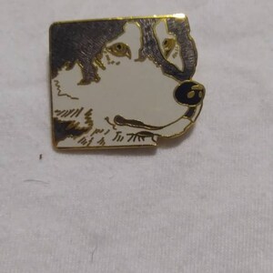 May include: A rectangular pin featuring a dog's head in profile. The dog has a white face with black and gray markings, gold trim, and a black nose. The pin has text at the bottom.