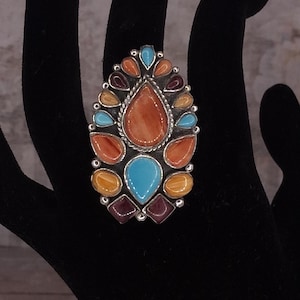 Op de afbeelding: Zilveren ring met een grote, druppelvormige oranje steen in het midden, omringd door kleinere stenen in blauw, oranje, geel en paars. De ring is geplaatst op een zwarte ondergrond.