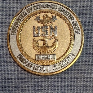 Puede incluir: Una moneda de oro y plata con las palabras "Presentada por el Jefe Maestro de Comando" y "CMDCM (SW) J. Clements" alrededor del borde. El centro de la moneda presenta un ancla dorada con un "USN" plateado encima.