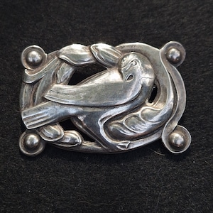 May include: A silver brooch featuring a stylised bird design with intricate details. The bird is surrounded by floral motifs and the brooch has four round accents.
