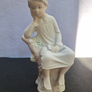 May include: A porcelain figurine of a young person sitting on a tree stump, holding a book and resting their chin on their hand. The figurine is painted in shades of white and beige.