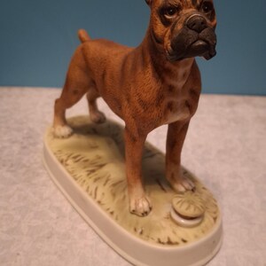 May include: A ceramic Boxer dog figurine, brown with a black muzzle and white paws, standing on a cream-coloured oval base. The base has a grassy texture and a small, round object. The dog is in a standing pose.
