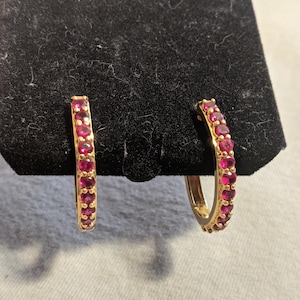 May include: A pair of gold hoop earrings featuring a continuous row of small, round, vibrant pinkish-red gemstones set along the front-facing curve of each hoop. The earrings are displayed side-by-side, showcasing the metallic gold finish and stones.