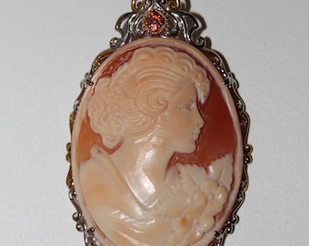 May include: Oval cameo pendant featuring a woman's profile carved in light peach and cream tones. The pendant is set in a silver and gold-toned frame, accented with small red gemstones. A classic jewelry piece.