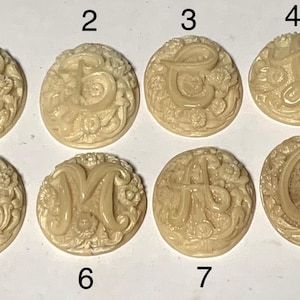 May include: Eight ivory-coloured buttons with raised letters and floral designs. The letters are A, B, C, D, E, F, G, and H.