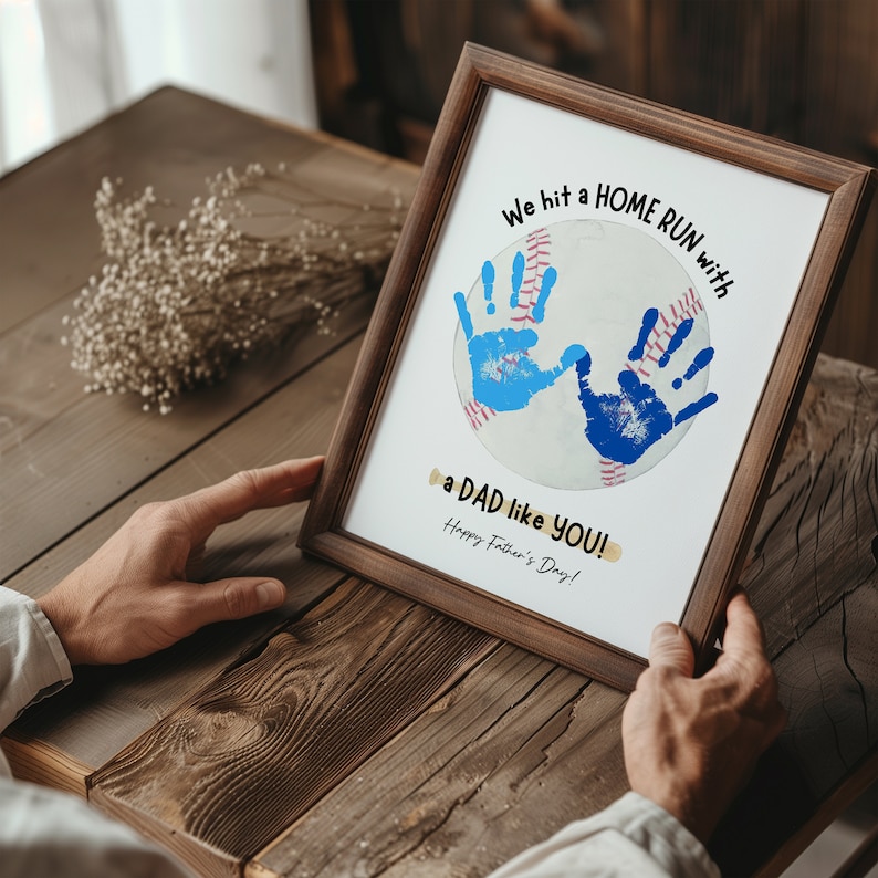 May include: A framed print with a baseball design. The baseball is decorated with blue handprints and the text "We hit a HOME RUN with a DAD like YOU! Happy Father's Day!"