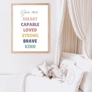 May include: A framed print with the text "You are SMART CAPABLE LOVED STRONG BRAVE KIND" in a white background with colorful text. The print is hanging on a white wall above a bed with white bedding and pillows.