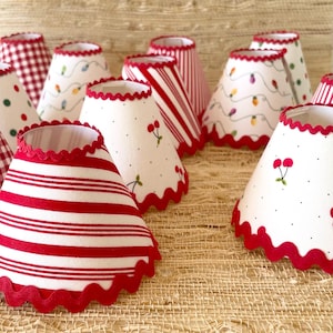 May include: A collection of cone-shaped lampshades with red scalloped edges. The shades feature various patterns, including red and white stripes, polka dots, and a string of lights. Some shades have a cherry design.