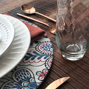 May include: A white dinner plate with a blue and red patterned placemat, two copper-toned forks, and a clear glass vase.
