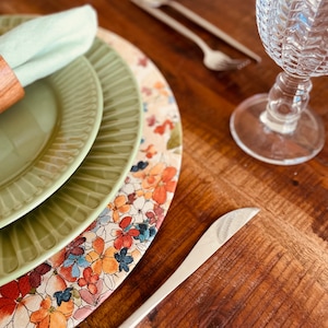 May include: A green and white floral charger plate with a green rim. The charger plate is layered on top of two green plates. A green napkin is rolled and placed on the top plate.