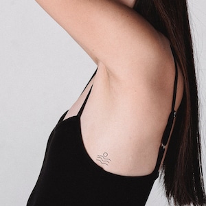 May include: A black tank top with thin straps. A small tattoo of a sun and waves is visible on the side. The person's arm is raised, and their face is partially obscured by their arm and long, dark hair.