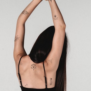 May include: A black tank top with thin straps. The arms are raised, displaying several small, black temporary tattoos. Designs include a moth, an eye, a leaf, a sunburst, a flower, and the word "MISSD".