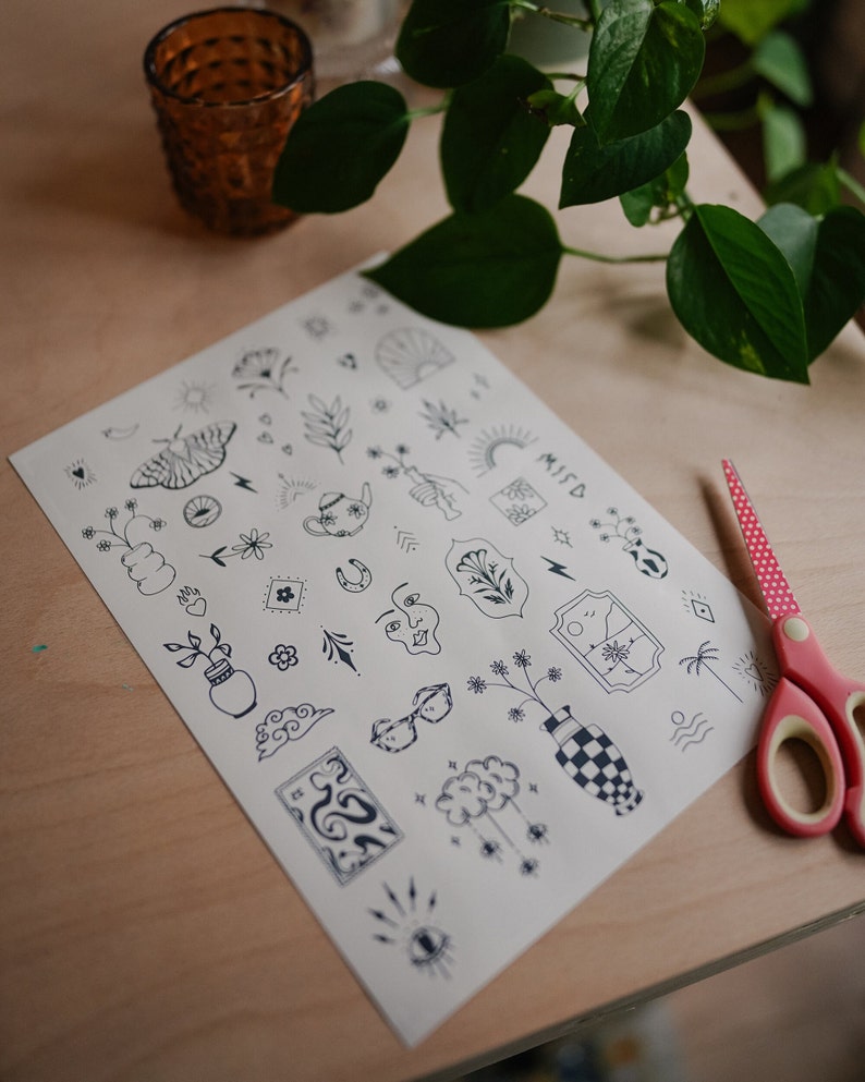 May include: A sheet of temporary tattoos with various black line art designs, including a butterfly, flowers, and clouds. The word "MISD" is visible. A pair of red and white polka dot scissors is on the right.