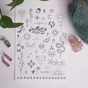 Könnte beinhalten: Eine Sammlung von Schmuck und Accessoires im Bohème-Stil auf einer weißen Oberfläche. Enthält ein Blatt temporärer Tattoos in Schwarz-Weiß mit Sonnen-, Mond- und Herzmotiven, eine kosmetische Tasche aus teal-farbenem Samt, eine rosa Haarspange und zwei Paar Ohrringe.