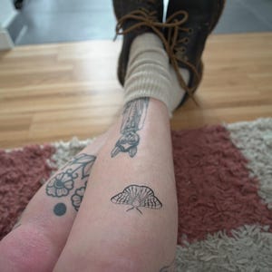 May include: A person's legs are crossed, with visible tattoos of a moth, owl, and floral designs. The person is wearing brown leather boots with tan laces and beige socks. The background includes a rug and wooden floor.