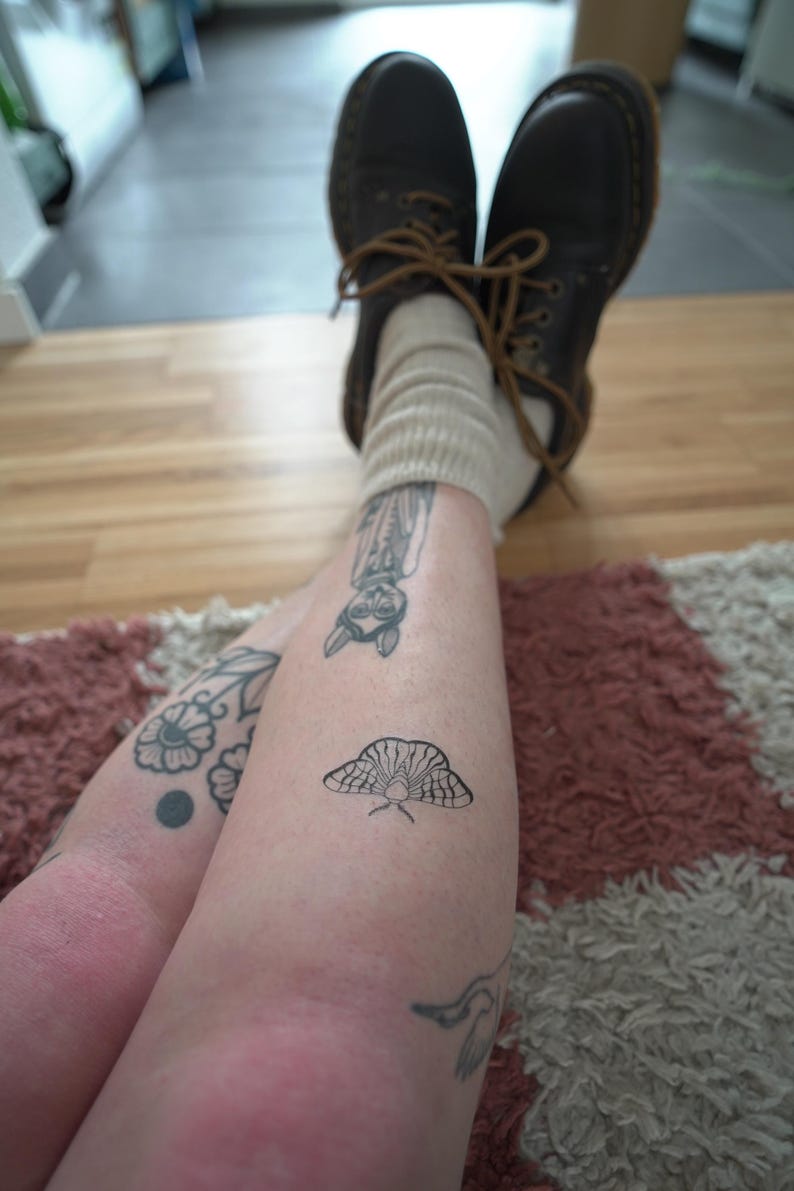 May include: A person's legs are crossed, with visible tattoos of a moth, owl, and floral designs. The person is wearing brown leather boots with tan laces and beige socks. The background includes a rug and wooden floor.