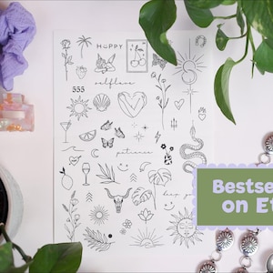 May include: A sheet of temporary tattoos featuring various black line art designs, including suns, hearts, butterflies, and botanical elements. The sheet is accompanied by a purple scrunchie, a decorative perfume bottle, and a silver bracelet. Text on the sheet includes "Happy" and "Selflove".