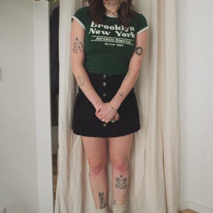 May include: A green t-shirt with white trim and the text "Brooklyn New York Athletic District Since 1997". The shirt is paired with a black button-down skirt, dark shoes, and a dark beanie. The person is wearing glasses and socks.