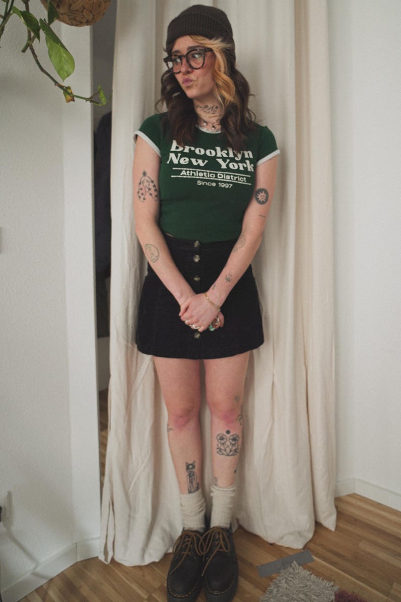 May include: A green t-shirt with white trim and the text "Brooklyn New York Athletic District Since 1997". The shirt is paired with a black button-down skirt, dark shoes, and a dark beanie. The person is wearing glasses and socks.