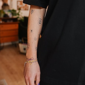 May include: A person's arm with several small black tattoos, including a leaf, a heart, and a face. A bracelet with yellow beads and a purple string is on the wrist. The person is wearing a black shirt.