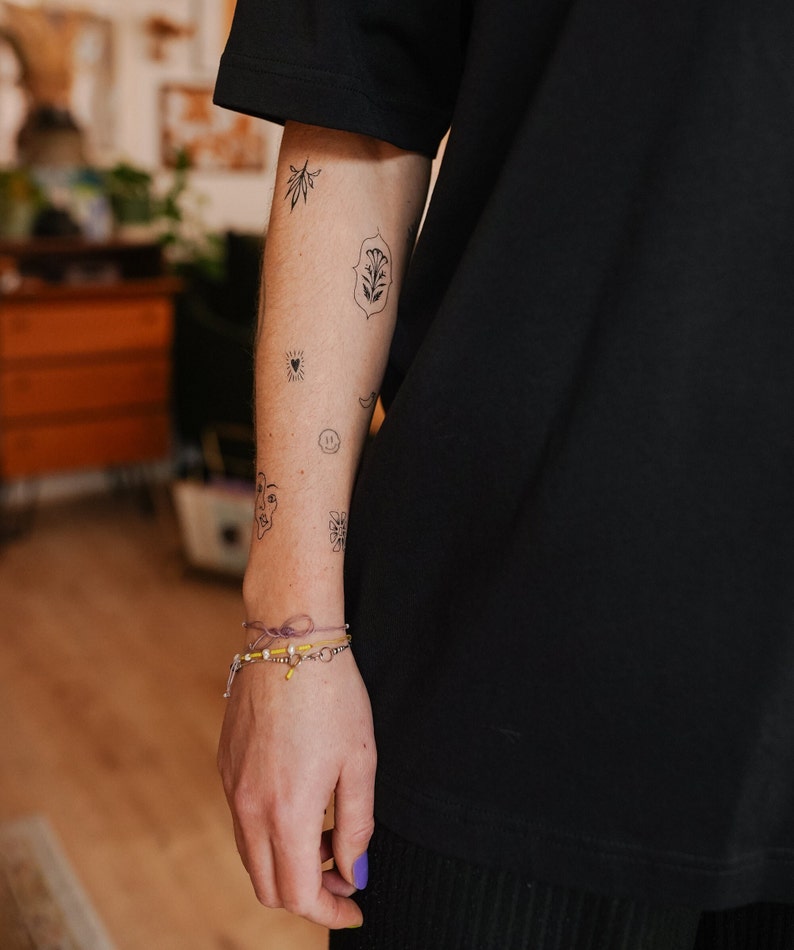 May include: A person's arm with several small black tattoos, including a leaf, a heart, and a face. A bracelet with yellow beads and a purple string is on the wrist. The person is wearing a black shirt.