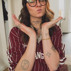 May include: A maroon and white striped long-sleeve shirt with the word "ANG" visible. The person is wearing a brown beanie, glasses, and has tattoos on their arms and neck.