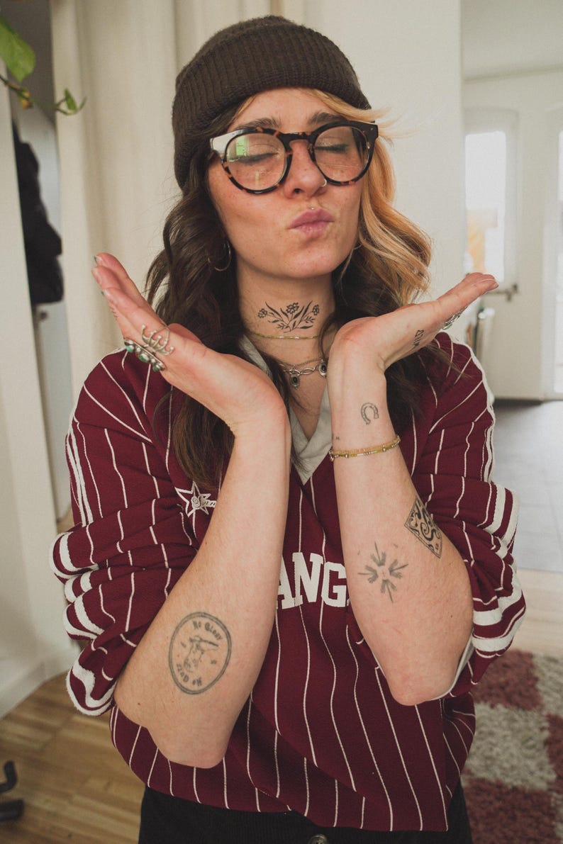 May include: A maroon and white striped long-sleeve shirt with the word "ANG" visible. The person is wearing a brown beanie, glasses, and has tattoos on their arms and neck.