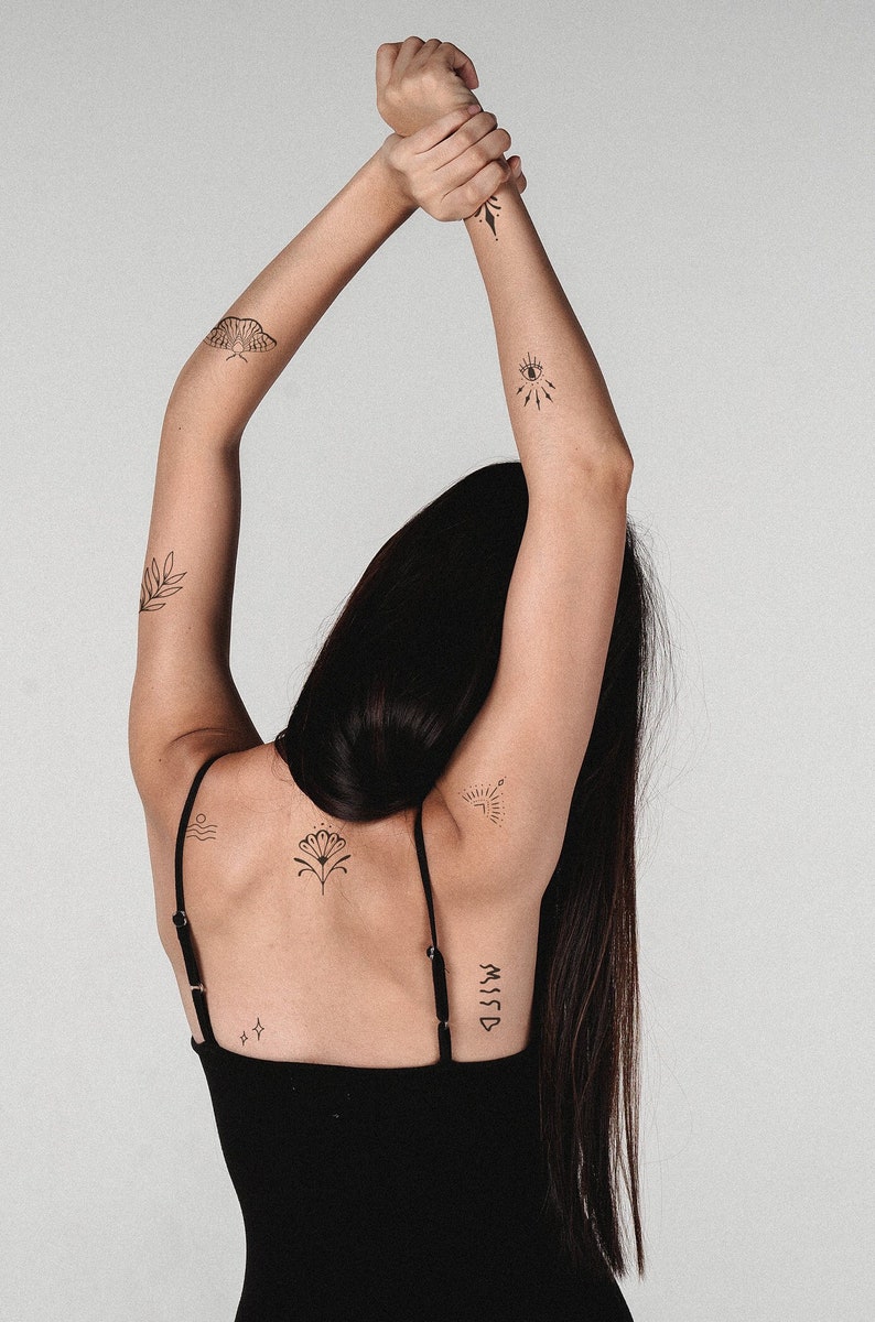 May include: A black tank top with thin straps. The arms are raised, displaying several small, black temporary tattoos. Designs include a moth, an eye, a leaf, a sunburst, a flower, and the word "MISSD".