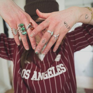 May include: A person wearing multiple rings with turquoise and amber stones, and a brown beanie. The person is wearing a maroon and white striped shirt with the text "OS ANGELES". The word "WILD" is tattooed on the hand.