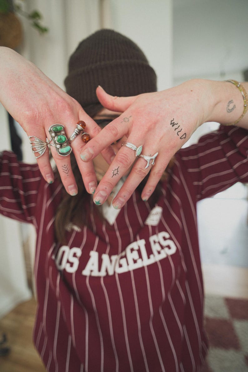 May include: A person wearing multiple rings with turquoise and amber stones, and a brown beanie. The person is wearing a maroon and white striped shirt with the text "OS ANGELES". The word "WILD" is tattooed on the hand.