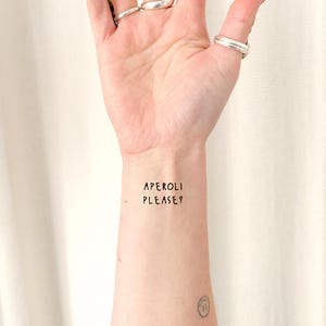 Könnte beinhalten: Ein temporäres Tattoo auf einem Handgelenk mit dem Text "APEROLI PLEASE?" in schwarzer, handschriftlicher Schrift. Der Hautton ist hell, und silberne Ringe schmücken die Finger. Ein kleines Smiley-Tattoo ist ebenfalls sichtbar.