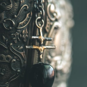 May include: A black heart-shaped pendant hangs from a silver chain. The pendant is attached to a cross-shaped charm with a detailed design. The pendant and chain are hanging against a dark, ornate background.