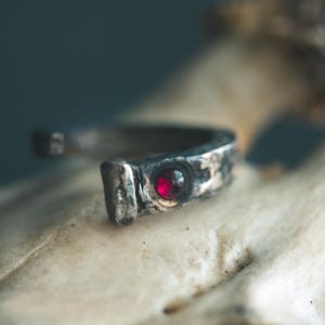 May include: A silver-toned ring with a hammered texture and a small, round, red gemstone. The ring has a unique, open design and is set against a blurred background.