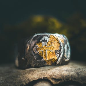 Puede incluir: Primer plano de un anillo de plata y oro con un diseño de cruz. El anillo tiene un aspecto texturizado y desgastado, con una cruz dorada en el centro. El anillo está sobre un fondo oscuro borroso.