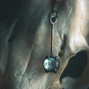 May include: A close-up of a silver-toned pendant with a light blue gemstone. The pendant hangs from a thin chain and is set against a blurred, off-white background. The gemstone is oval-shaped and set in a dark metal setting.