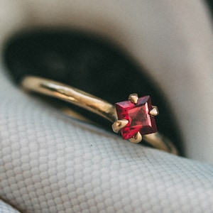 May include: A gold ring with a square-shaped red gemstone set in a prong setting.