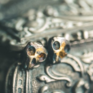 May include: Two small silver skull-shaped earrings with gold accents. The earrings are sitting on a textured silver surface.