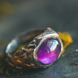 May include: A ring featuring a large, smooth, oval-shaped purple gemstone set into a dark, textured metal band. The metal surface has a rugged, uneven appearance with deep grooves and a matte finish, contrasting with the polished, reflective purple stone.