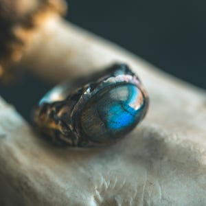 May include: A ring featuring an oval-shaped labradorite gemstone with a prominent blue flash. The band is crafted from dark, textured metal with an uneven, organic surface that surrounds the stone in a bezel setting, creating a rugged appearance.