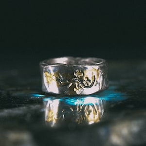 May include: A silver ring with a gold design. The ring is on a dark blue surface.