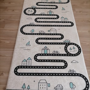 Puede incluir: Una alfombra de juego beige con un camino negro que serpentea a través de una ciudad con casas, árboles, coches y bicicletas. El camino está delimitado por una línea discontinua.