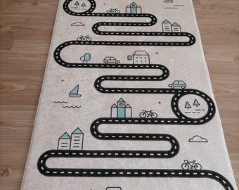May include: A beige play mat with a black road winding through a town with houses, trees, cars, and bicycles. The road is outlined with a dashed line.