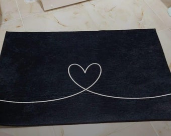 May include: Black bath mat with a white heart design. The heart is formed by a continuous line.