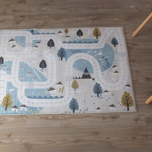 Puede incluir: Una alfombra de juego azul claro y blanca con dise&ntilde;os de carretera y animales. La alfombra presenta un camino sinuoso con &aacute;rboles, monta&ntilde;as y animales en el camino.