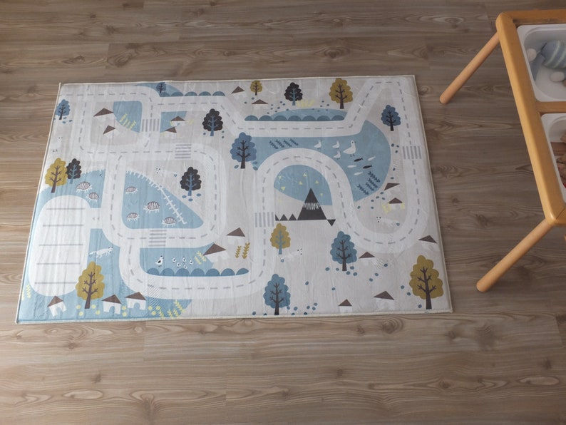 Puede incluir: Una alfombra de juego azul claro y blanca con dise&ntilde;os de carretera y animales. La alfombra presenta un camino sinuoso con &aacute;rboles, monta&ntilde;as y animales en el camino.